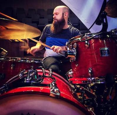 Justin is seen playing a red drum kit from the perspective of his kick drum.